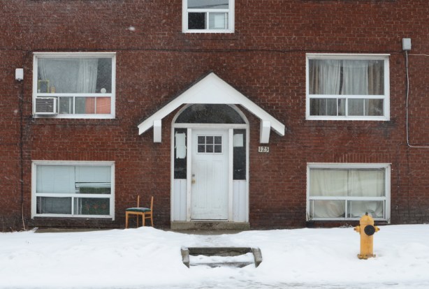 side of a brick multi family residence, windows, white door, with a small white porch over the door, broke chair beside the door