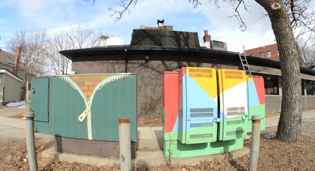 street art on two metal boxes on the sidewalk, one is a zipper opening to reveal a brick wall 