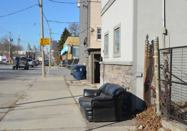 an old black vinyl couch on a sidewalk on Eastern Avenue, beside a house 