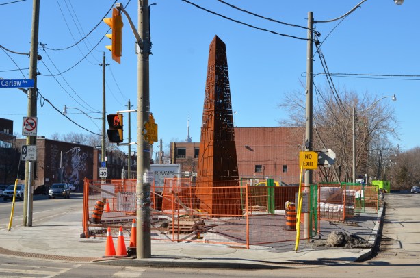 a new public art installation at Carlaw and Dundas with construction fence still around it, also a lot of utility poles around it, rusted metal cut with lasers in a design, sculpture is the shape of an obelisk 