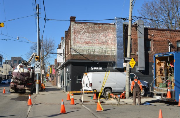 workmen at an intersection, white van parked, in front of building with ghost sign for Coady sweets, new Spanish restaurant on the corner 