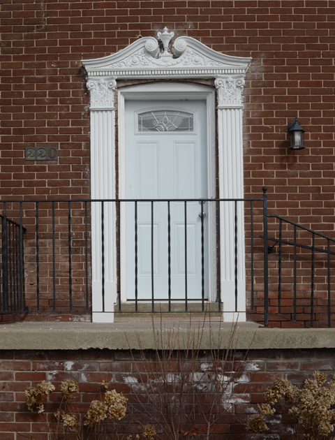 white door on small porch with black railing. door has fancy white trim with details on top 