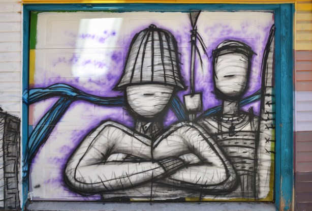 street art on garage door, faceless man with hat on and arms folded, behind hime another faceless person, all are black line drawings, with some shading.