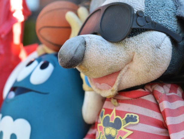 an old stuffed mouse with black plastic glasses, in front of a blue m & m character throwing a basketball, outside, and slightly weathered 