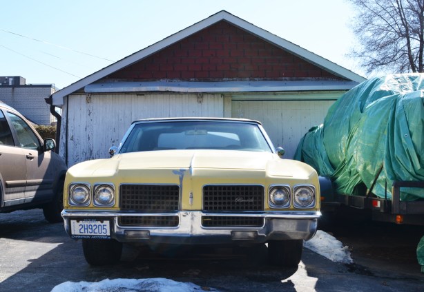 an old yellow Oldsmobile car, with historic licence plate, parked in a driveway in front of an old white garage 