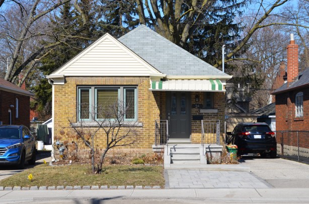 a well kept yellow brick post war bungalow with a grey roof and a partial white and green metal awning over the front steps that lead to a small porch 