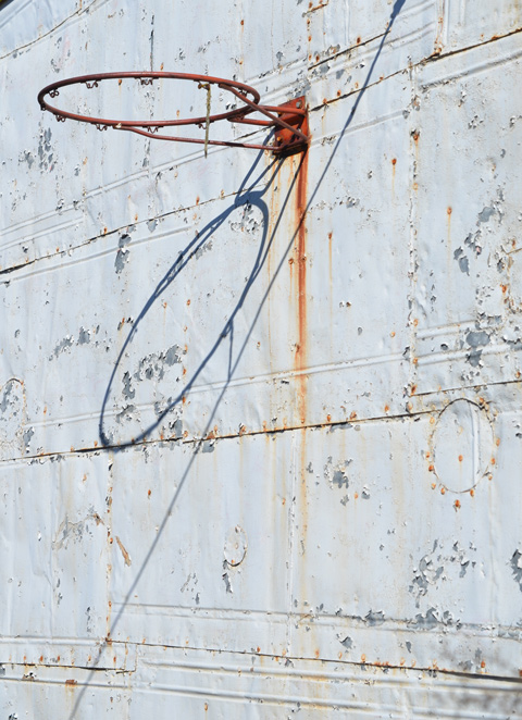 metal wall, painted white, exterior, with some rust, also an old basketball hoop and its shadow 