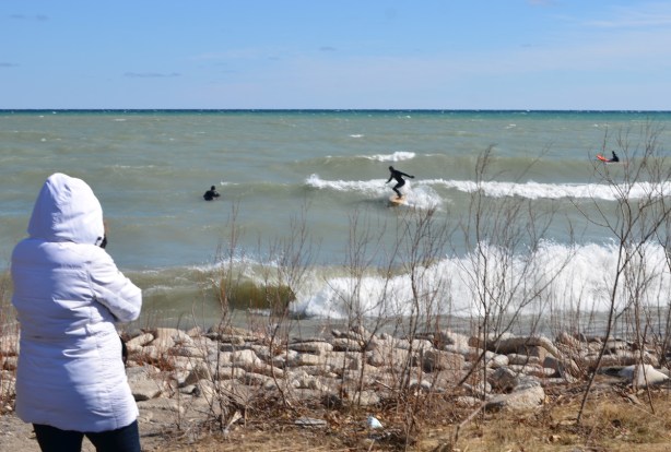 a woman in a white parka is watching men surf in rough waters and high waves of Lake Ontario 