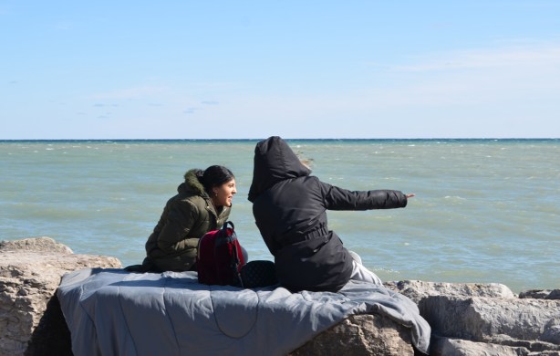 two women in winter clothes sitting on a blanket on rocks by the lake, one is pointing at something