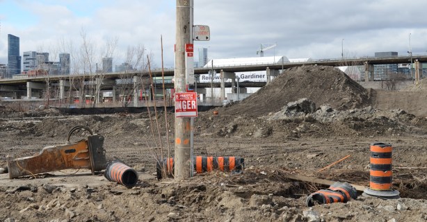TTC bus stop for route 72 on Commissioners street but pole is now in the middle of a construction site, lots of dirt, orange and black traffic cones, Gardiner Expressway in the background