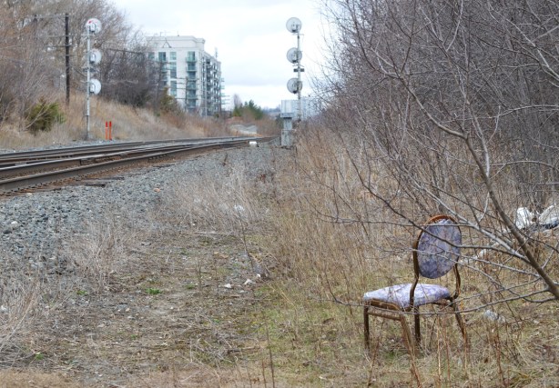 a lone chair sitting in the grass beside the railway tracks, shrubs behind the chair, early spring, no leaves on the shrubs 