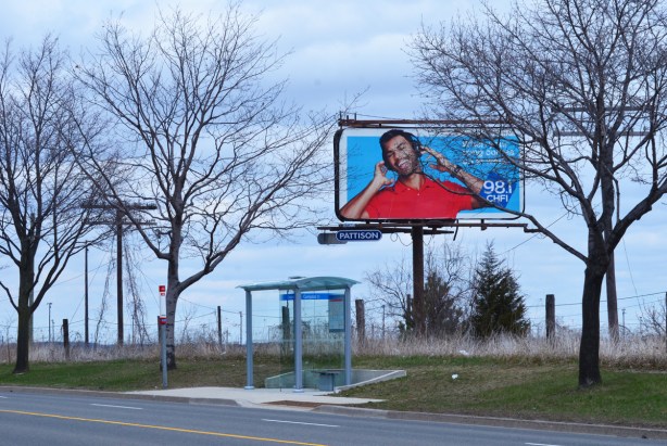 a bus shelter on the side of a street, with vacant land behind and a large billboard advertising a radio station