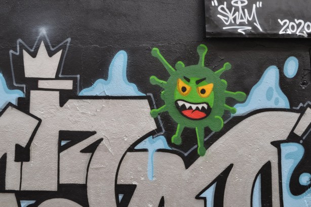 part of a mural in Graffiti Alley, a silver tag with a green corona virus with open mouth, white teeth and yellow eyes