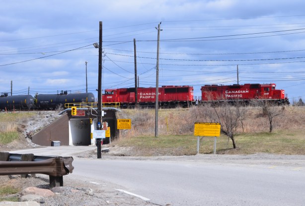 two bright red CPR engines at the front of a train, black tanker cars behind, as it crosses over the bridge at the entrance to the CPR railway yard