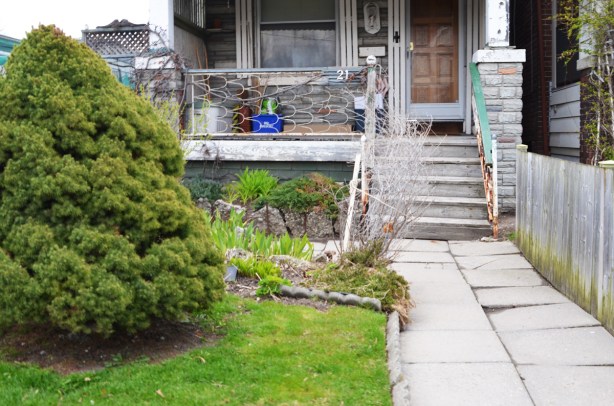 front yard and porch of a house, walkway is concrete slabs that are uneven, pine bush on grass, metal railing on porch, small garden in front of porch 
