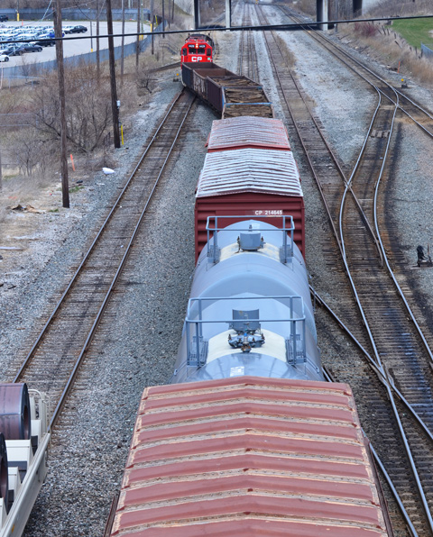 seen from a bridge, a train passes below, engine, flatbed cars, a tanker, and a boxcar 