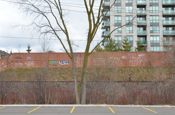 edge of parking lot that it empty, with railway tracks behind, a wall with graffiti, and an apartment building in the background 