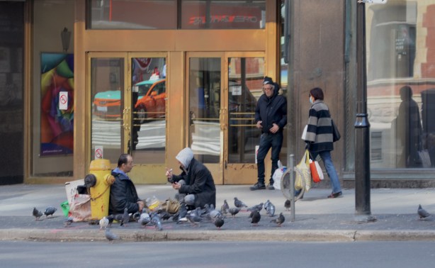 two men sitting on the sidewalk feeding pigeons, many pigeons, a security guard stands by a door behind them and a woman with a face mask walks past 