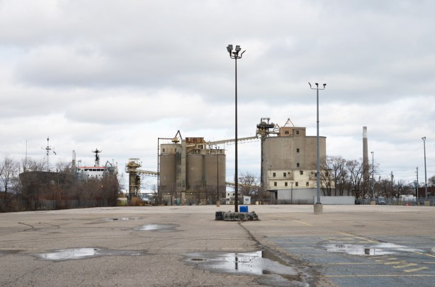 large empty parking lot in the foregraound, largare cement facitily in the background, with a large laker docked beside it 