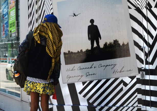 a woman in a yellow and black striped scarf stops to look at a picture on a wall decorated with many black and white stripes going in many different directions, on the stripes is an enlargement of a polaroid picture of a man standing in a field with an airplane flying over him