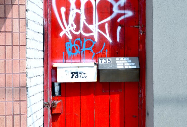 bright red gate between two buildings, 2 mailboxes on the gate, one white and the other a brass colour. Brass mailbox is 735