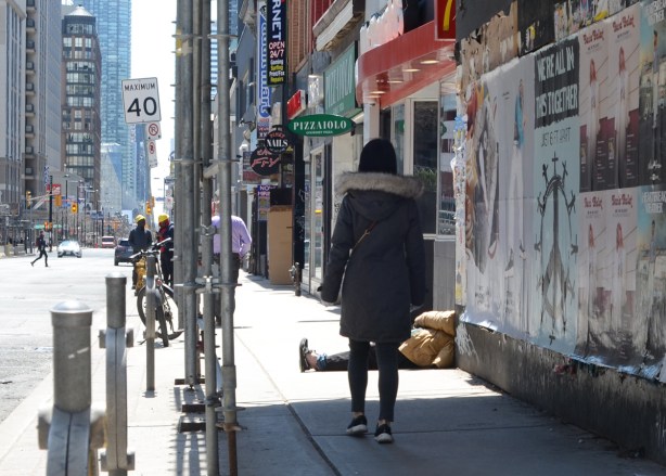 a woman walks down Yonge Street under a covered walkway (for construction) and towards a man half sitting and half lying on the sidewalk, with one leg stuck out into the sidewalk 