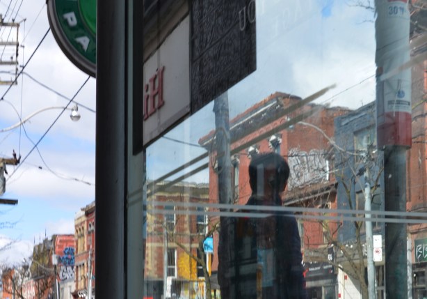 a person waiting beside a bus shelter on Queen West, seen from the back including reflections in the glass of the shelter 