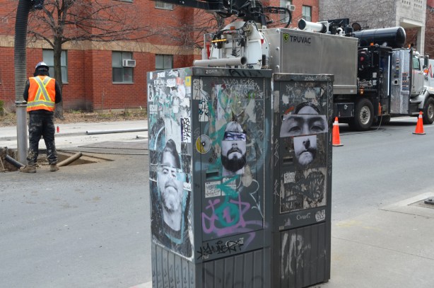 paper paste up graffiti of faces and eyes by Jeremy Lynch on metal boxes on the sidewalk, with construction workmen in the background 