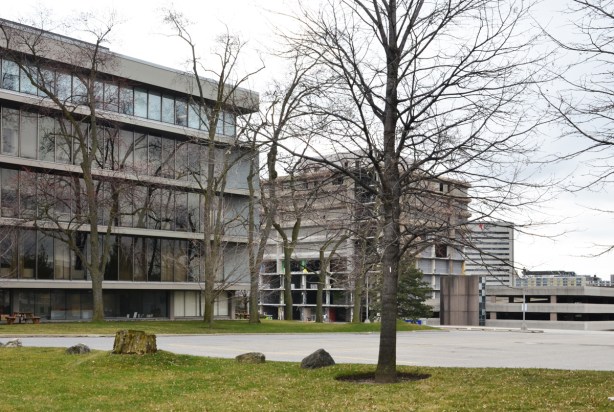 two concrete buildings from the 1970s or 1980s, one behind has started to be demolished, a parking lot and large tree between the buildings