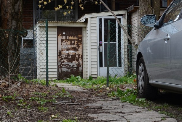 back of a house, silver car parked, patio stone walkway to back door. screen door as well as old mottled brown and beige door, small stairs to back porchwhere there is a white chair 