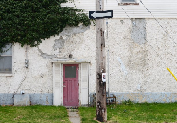 faded red, now pink, door on a white house, dirty and greyish stucco on the exterior, small bit of grass in front, one way sign on the utility pole in front of the house. 