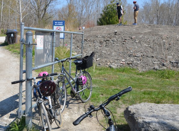 bikes parked against a fence with signs on it, with graffiti words that say flow like a river, in the background a mound of dirt with two young men standing on top of it. 