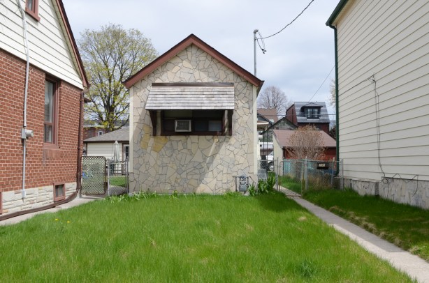 very small beige house with one window in the front, large grassy front yard, between two largeer houses that are closer to the street 