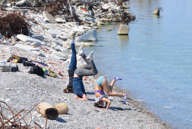 three young people trying to do headstones on the pebbles at the shore beside Lake Ontario, Tommy Thompson Park, with old rusty rebar in piles on the shore two, washed up old trees and roots,