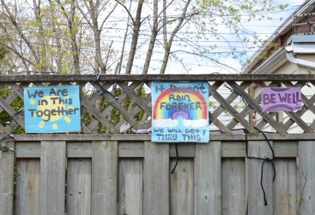 three small painted signs mounted on a wood fence, one says be well, another is a rainbow and the third says we're all in this together