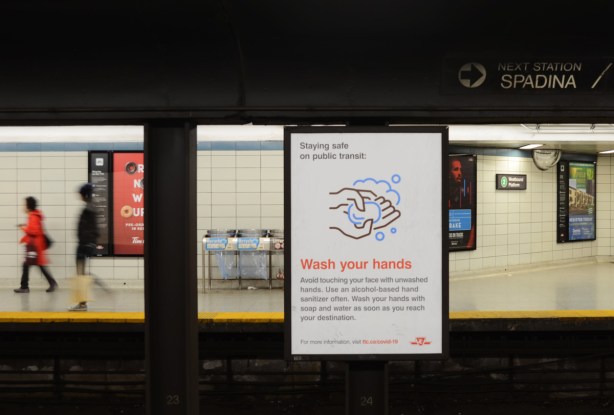 subway station platform, a couple of people walking, a sign reminding people to wash hands frequently and thoroughly because of covifd19