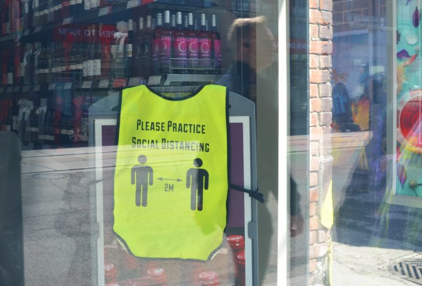 looking in the window of a wine store. A yellow vest is hanging by the counter, with words on vest that say please practice social distancing, two silhouette people 6 feet apart 