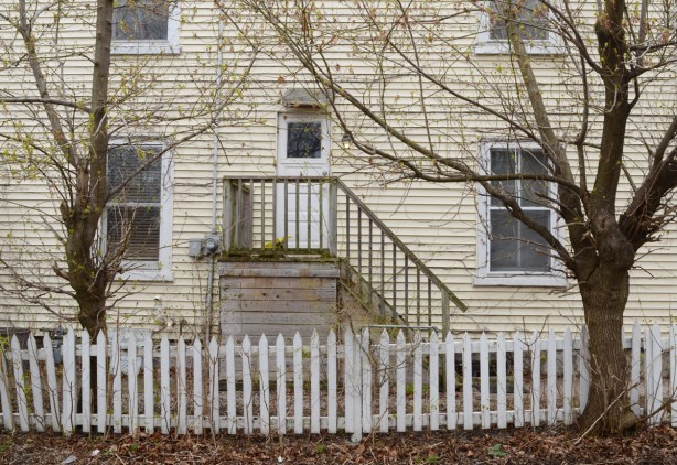 white picket fence along the side of a beige house with two large trees in yard, a door with newer wood porch and steps 
