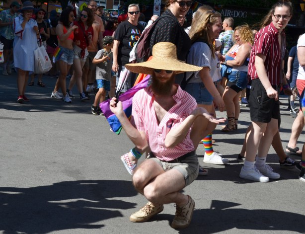 man in pink shirt and straw hat dances while squating while people passing by slow down to stare 