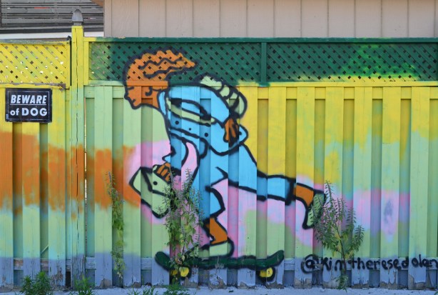 mural on a yellow fence of a person on a skateboard with a bag in one hand, by kim therese dolan 
