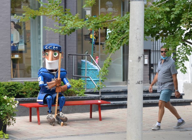 a metal statue of a hockey player in Toronto Maple Leaf blue sits on a bench outside a gallery, wearing a covid face mask, as a man walks past 