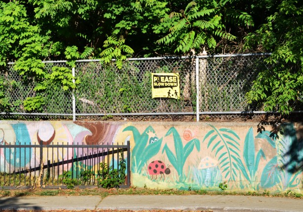 a sign that says slow down on a fence above a concrete wall with paintings of plants and flowers on it