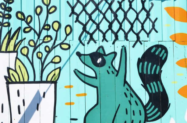 teal raccoon in a mural looking at a plant growing in a white planter 
