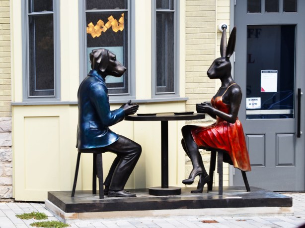 male dog in blue and female rabbit in red sitting face to face at a table with coffee, sculpture on Scollard street