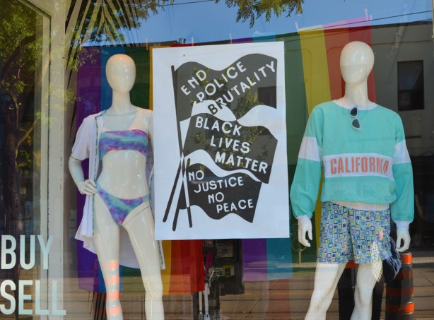 store window, two mannequins in summer attire, woman in bikini and man in shorts and long sleeve t-shirt, also a black lives matter poster