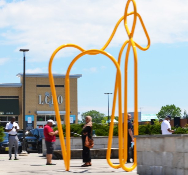 large, tall, sculpture made of yellow tubular metal in the outline shape of a horse in a mall parking lot with people standing around and under it