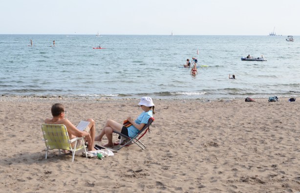 a couple sit on low chairs in the sand at Woodbine Beach, others are in the water swimming or on paddle boards 