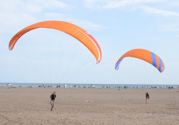 two men with large kites are trying to get themselves up in the air, a woman in a bathing suit lies on the sand between them. 