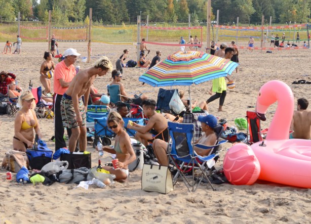 a group of young people in bathing suits and summer clothing having a picnic at the beach, some under a striped umbrella and some standing and sitting around an large inflatable pink flamingo 