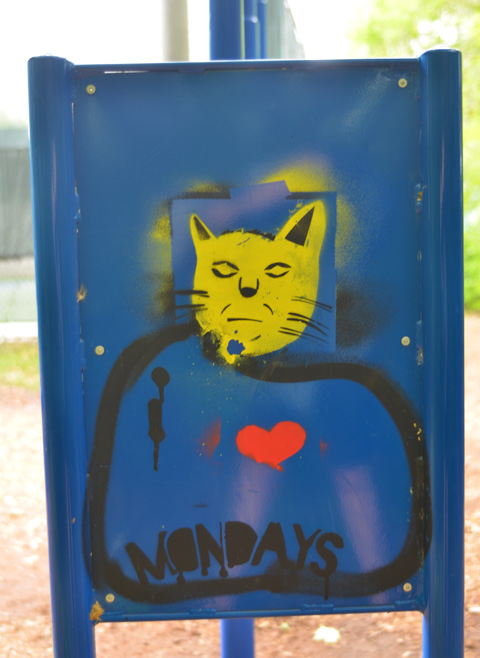 street art on the back of a blue metal sign, a yellow cat head and the words I Mondays, with a red heart between I and Monday, therefore I love Mondays